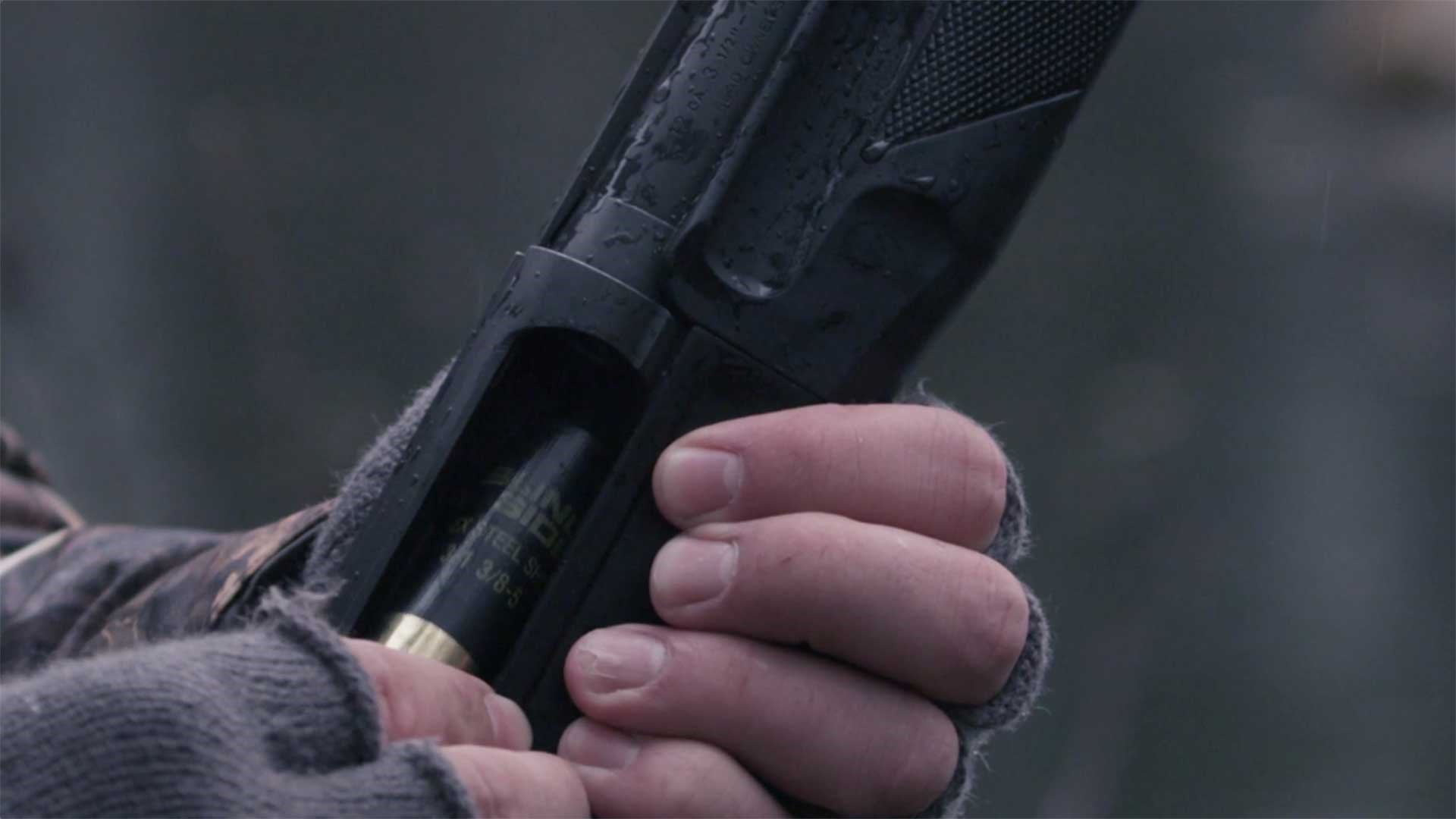 A man with gloved hands inserts a shotshell into a black hunting shotgun.