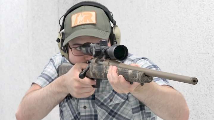 Man wearing ballcap and protective gear is holding a rifle on a shooting range.