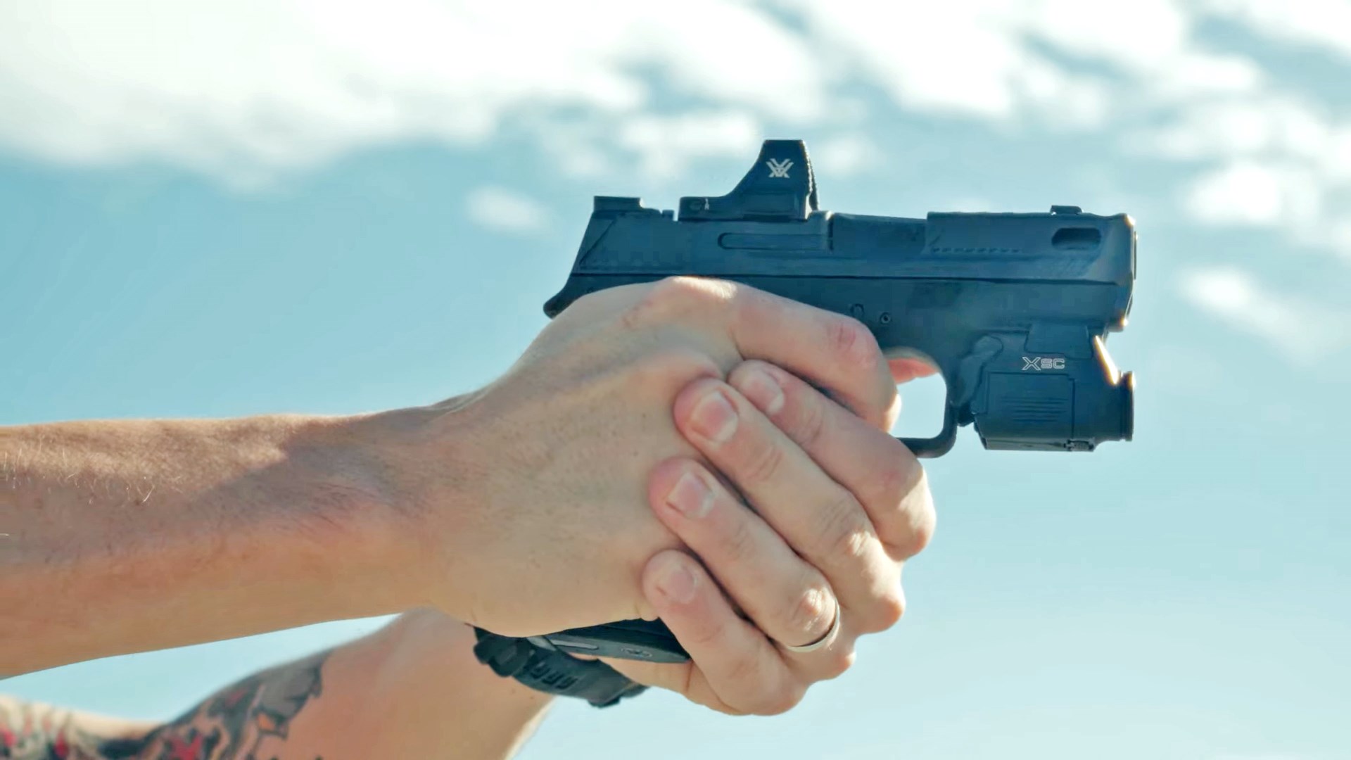 Man aiming a Smith & Wesson Shield X Carry Comp in front of a cloudy, blue sky.
