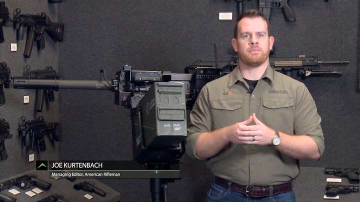 Man standing with hands crossed in gray room with guns on walls.