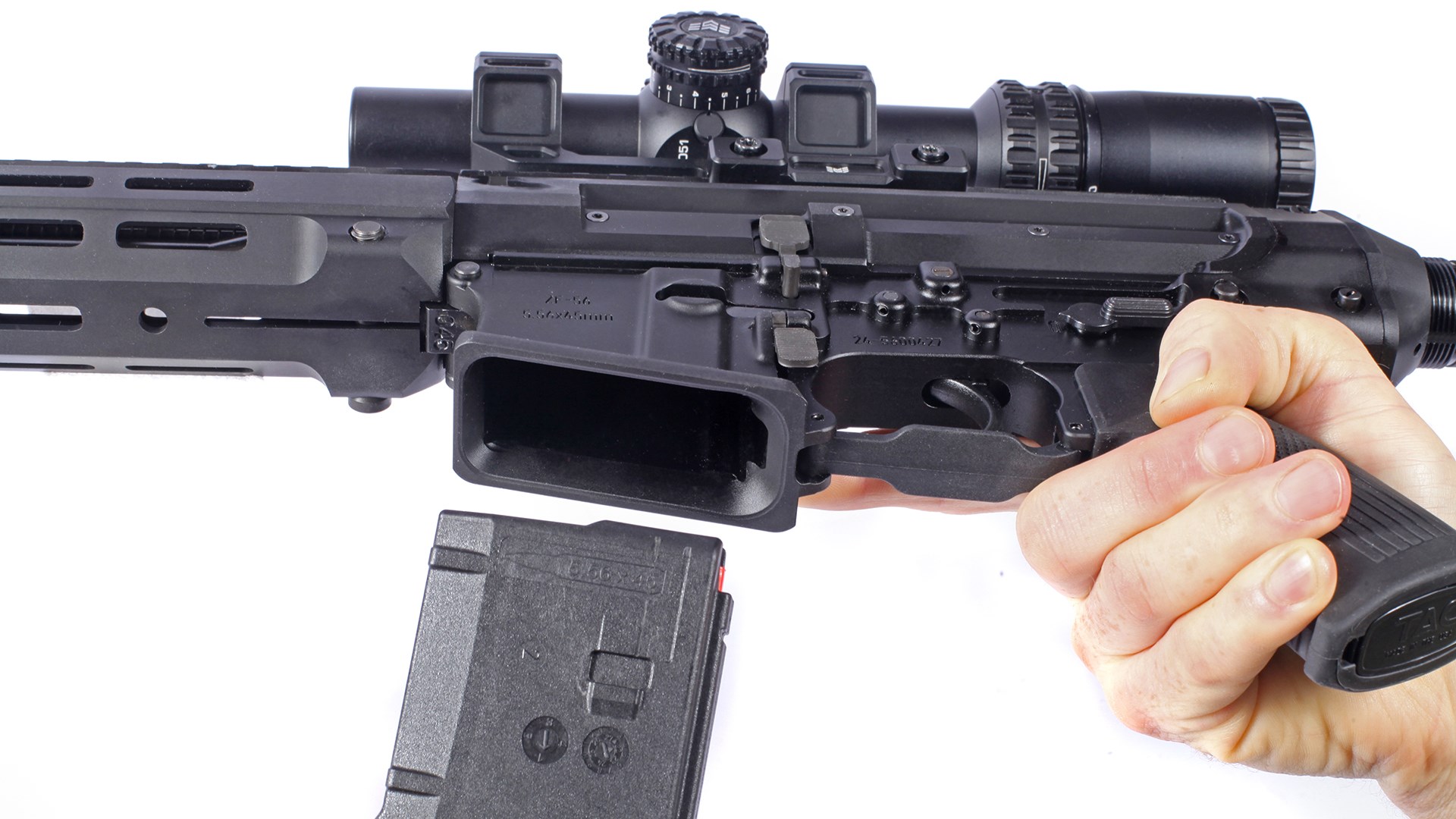 Zenith Firearms ZF-56 rifle in hand left-side, bottom-side view with magazine well open, hand holding and magazine present near well shown on white background with riflescope attached