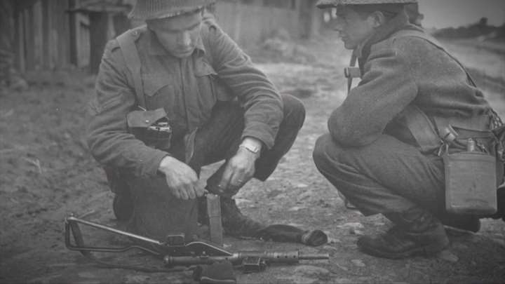 World War II British soldiers peering down at a Sten gun in a black and white image.