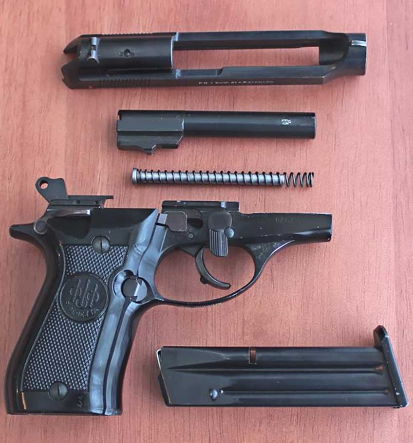A disassembled Beretta Model 81 on table with slide on top, barrel and guide rod in middle and pistol frame below with magazine.