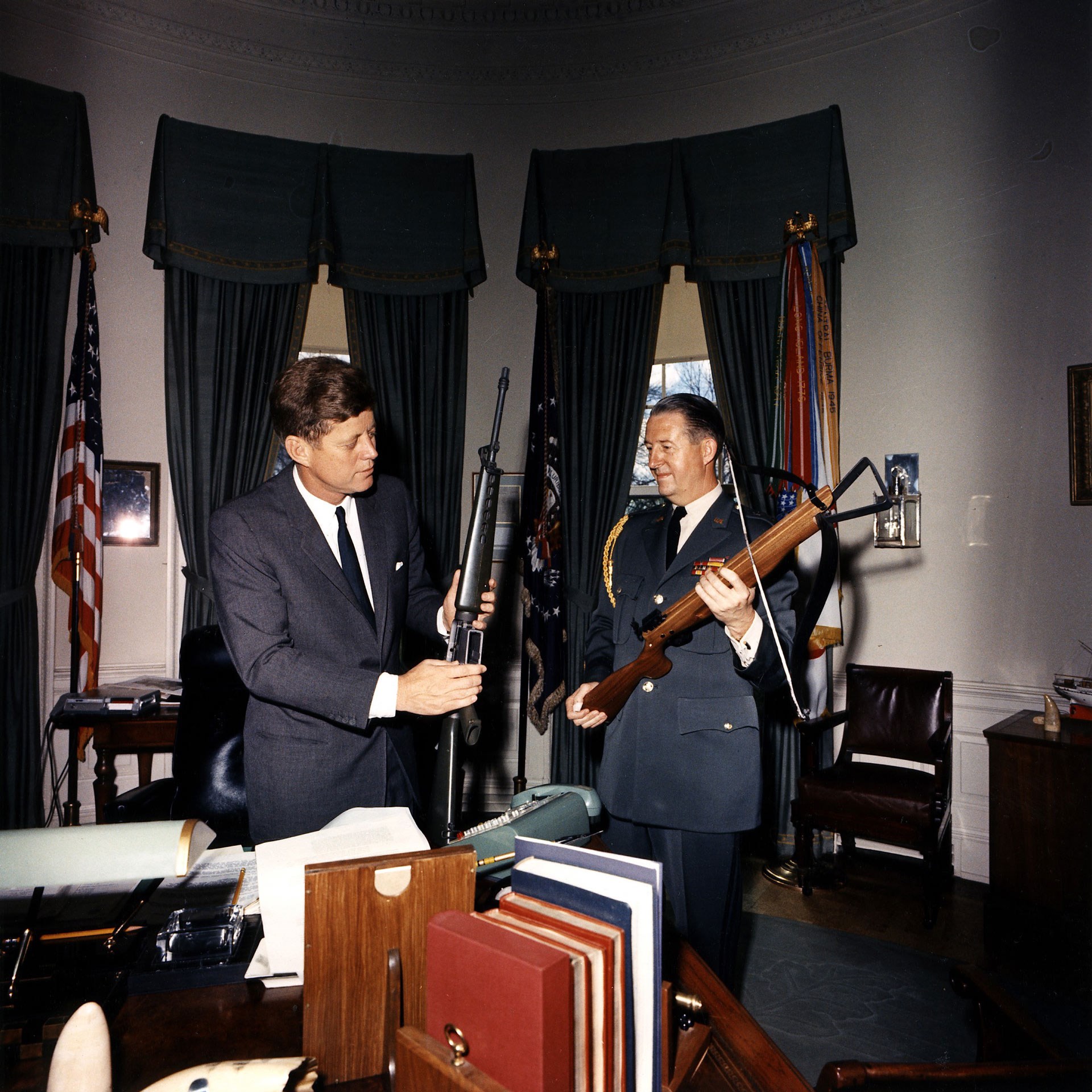 President John F. Kennedy and Military Aide Gen. Chester V. Clifton examine a COLT Model 601 rifle and crossbow in the White House’s Oval Office on April 19, 1963.