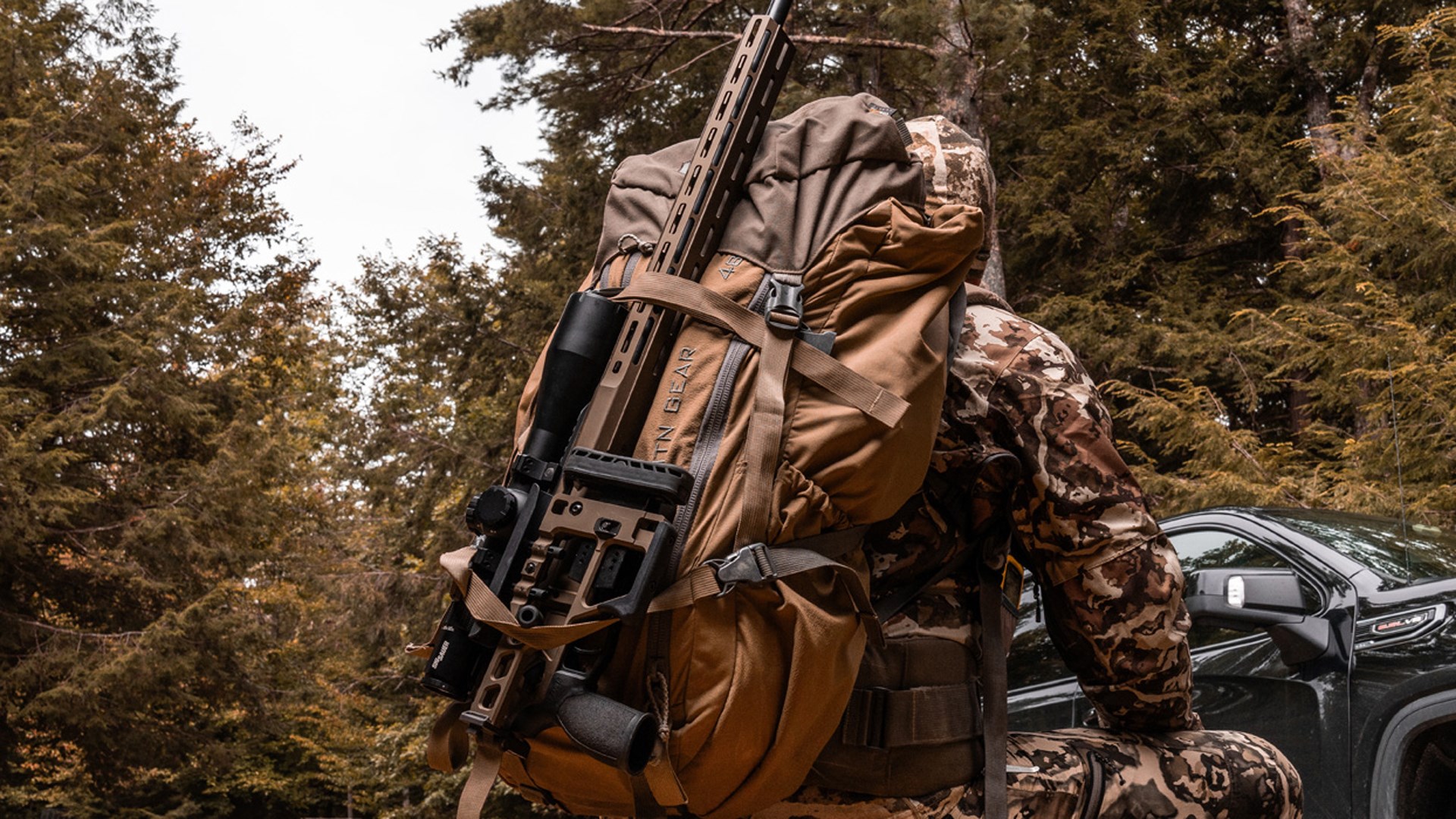 A SIG Sauer Cross Magnum is stowed on a hunter's backpack as he treks through the woods toward a black truck.