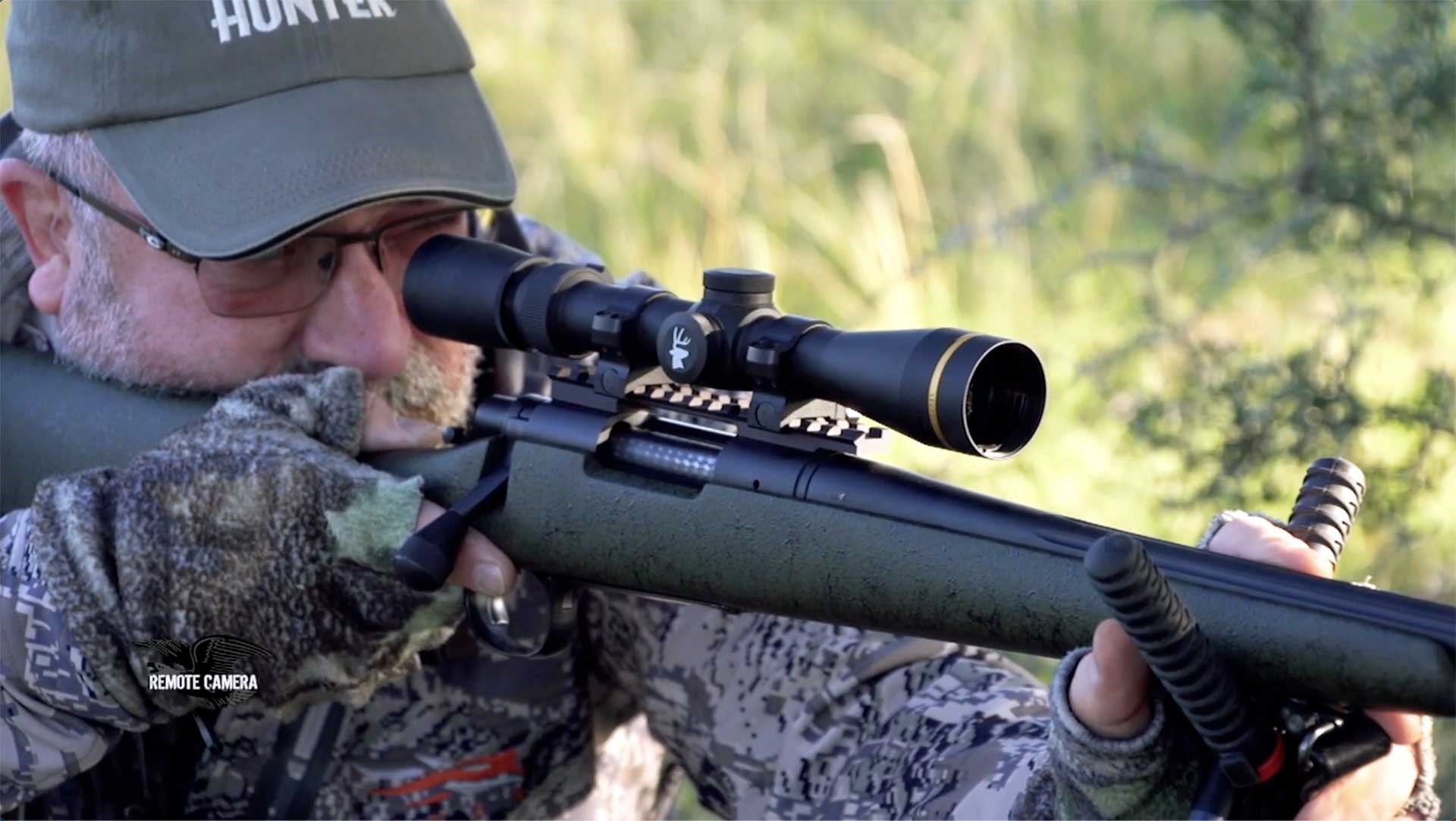 A man aims a hunting rifle off shooting sticks in the woods.