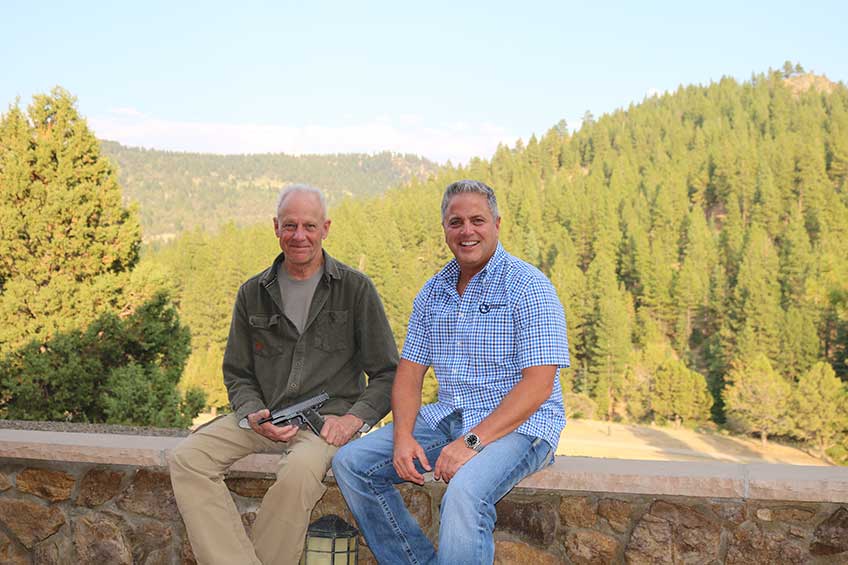 Clint Smith of Thunder Ranch and Mark Stone of Nighthawk Custom sit on a stone ledge with green pine forest and blue sky in the background.