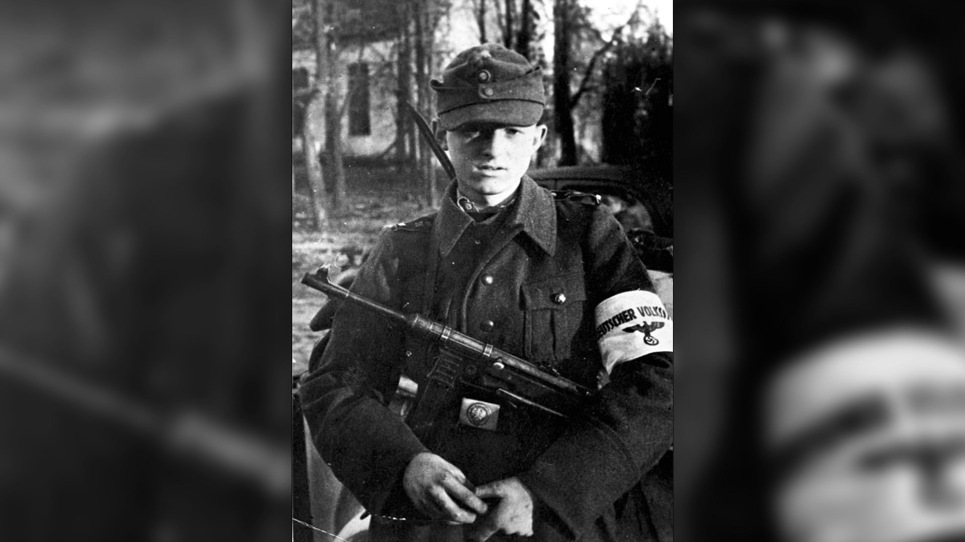 Image overlay from 1945 of young German Volkssturm in uniform with white patch on arm slung MP40 submachine gun over shoulder in front of church
