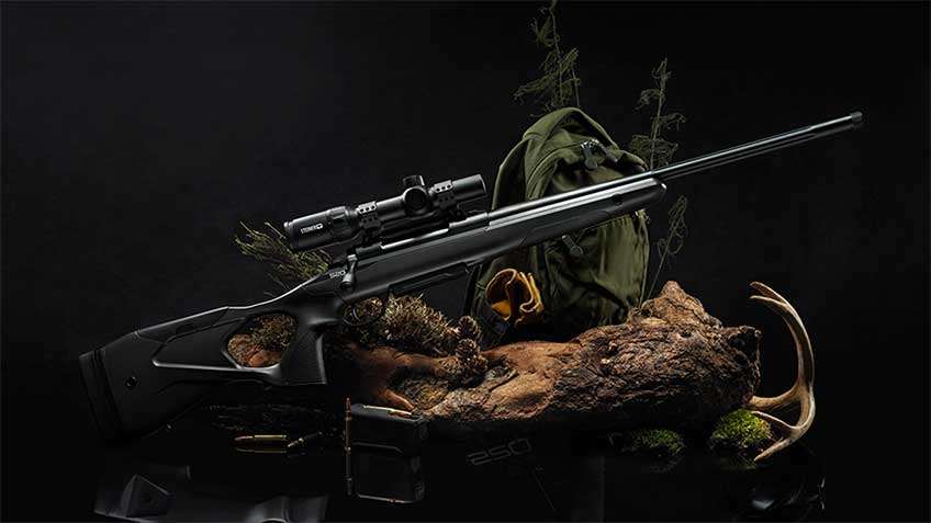 Right-side view Sako S20 bolt-action rifle with scope attached shown with dark outdoor scene and backpack.