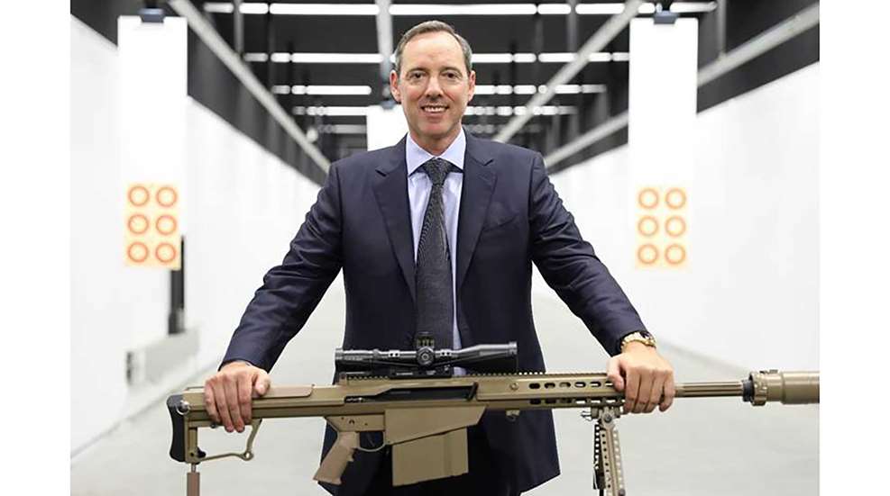 NIOA Group CEO Robert Nioa stands on a bright-white indoor shooting range, resting his hands on a tan Barrett .50-caliber semi-automatic rifle.