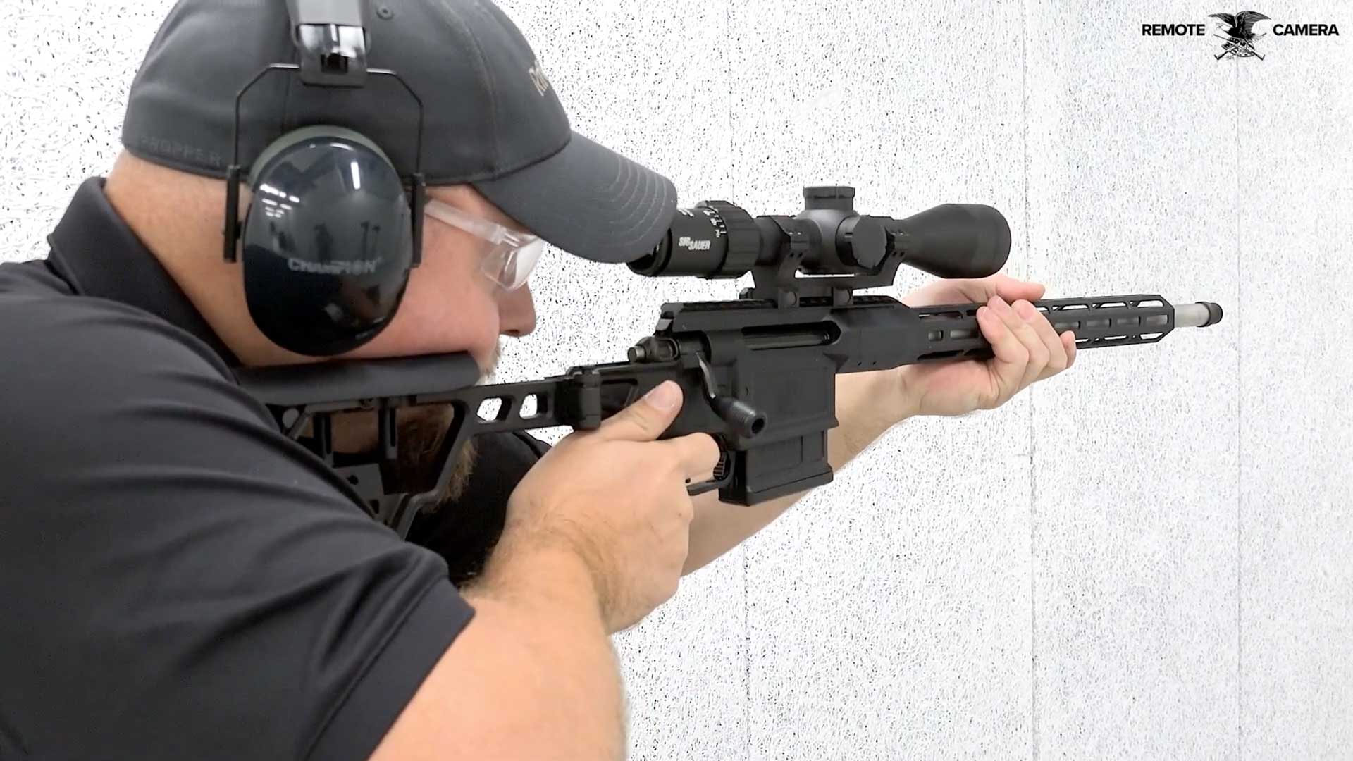 man wearing black shirt ballcap and earmuff shooting black bolt-action rifle from SIG Sauer with text on image noting "REMOTE CAMERA"