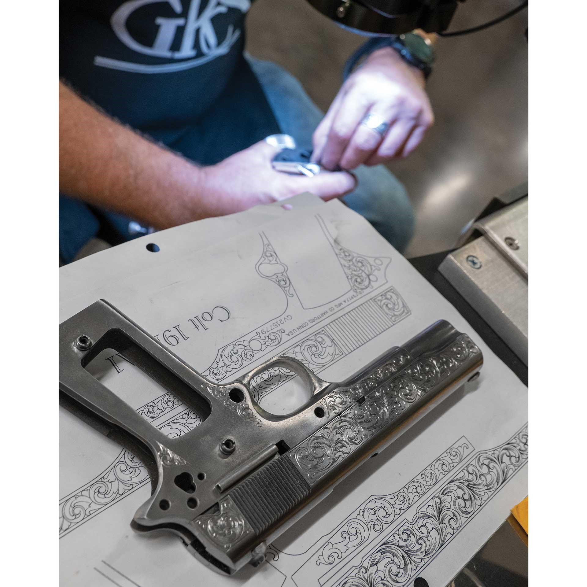 A man works to engrave a firearm while looking at a pattern in the foreground.