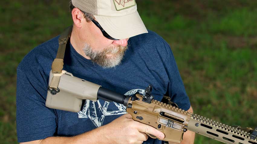 man wearing ballcap and blue shirt examining rifle ar-15 gun outdoors grass