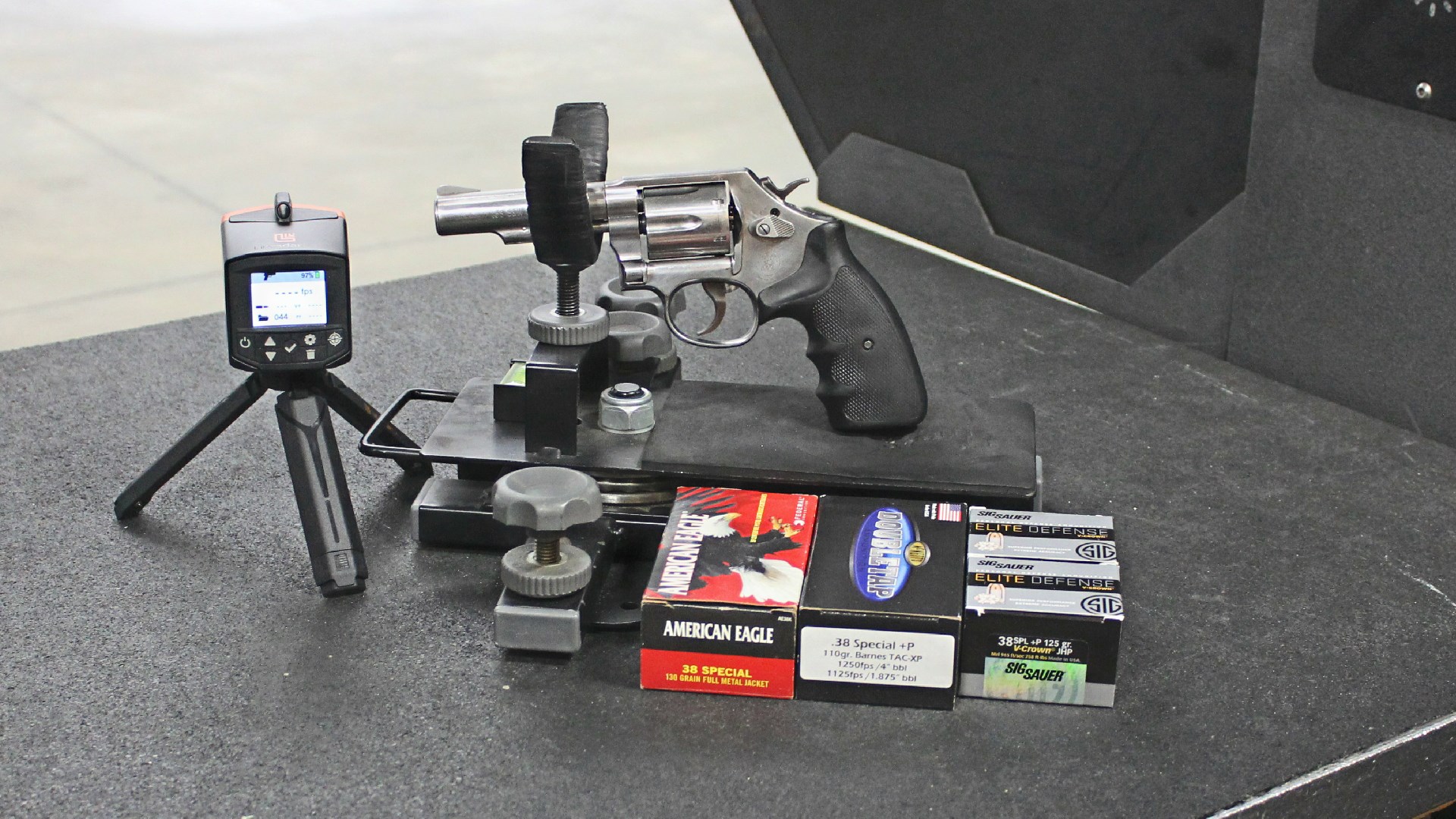 Smith & Wesson Model 64 stainless steel revolver with black rubber stocks in cradle shooting rest on range with boxes of ammunition and LabRadar LX chronograph on tripod left side