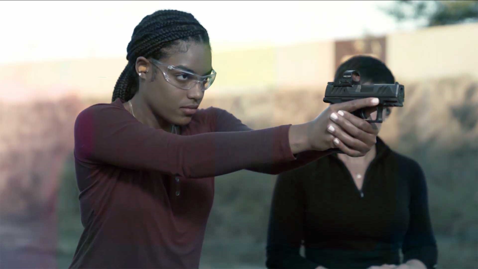 A woman aims a Walther handgun downrange while an instructor watches in the background.