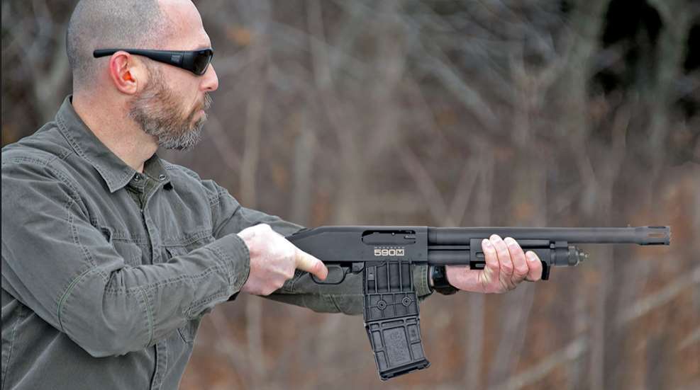 Bald bearded man wearing sunglasses firing in hand new Mossberg pump-action shotgun right-side view with detachable box magazine outdoors
