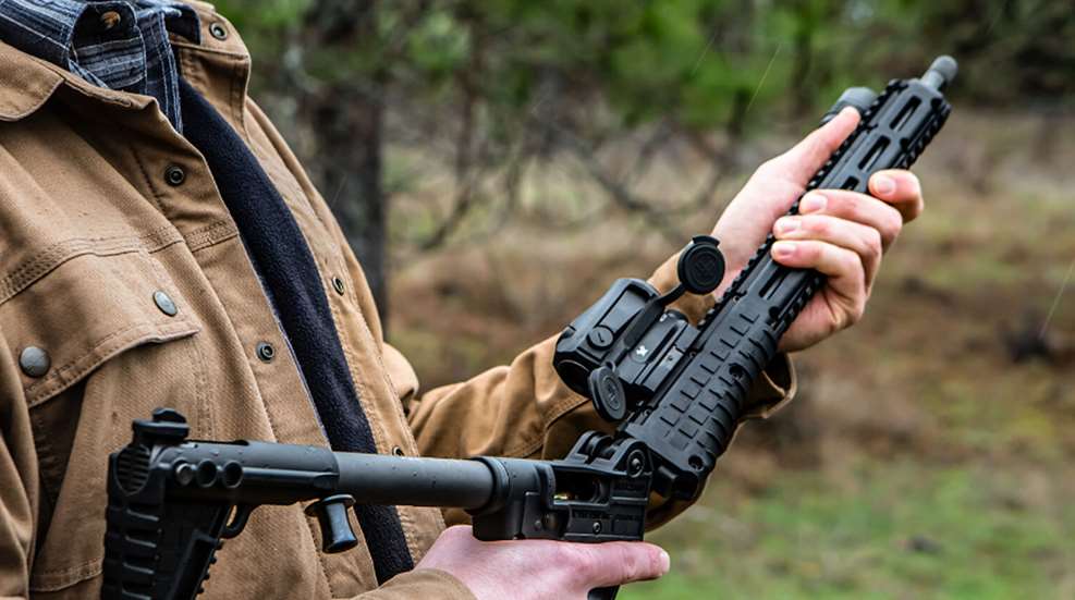 KelTec SUB2000 GEN3 folding pistol-caliber carbine shown outdoors in the hand unlocking the folding mechanism grass trees soft focus background man watching.