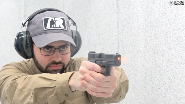 Beared man with glasses, hearing protection and gray ballcap shooting a Glock G44 rimfire pistol on white shooting range. Photograph shows fire emitting from barrel of pistol.