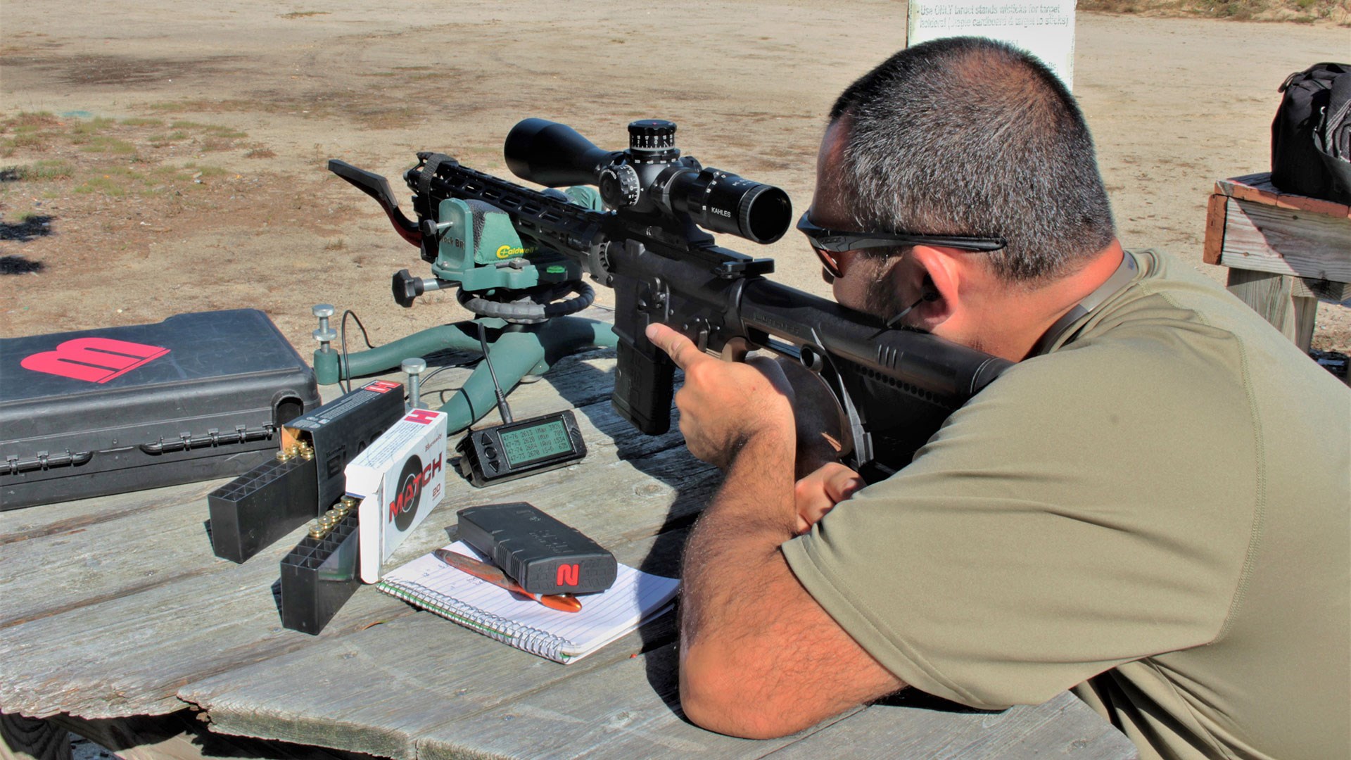 man shooting rifle outdoors with gear on table